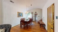This single-floor space exudes a distinctive Spanish Colonial character, blending classic architectural details with warm, inviting interiors. Rich, dark hardwood floors run throughout, anchoring each room with a sense of depth and tradition. The main living area is a generously proportioned room, highlighted by striking teal-framed French doors that open directly onto a lush, plant-filled courtyard. A crystal chandelier adorns the ceiling, adding an elegant focal point, while built-in white shelving lines one wall, offering functional display space. Natural light streams in through the French doors, illuminating the open interior. A hallway with a dramatic barrel-vaulted ceiling connects the living areas, lending an airy, cathedral-like quality to the passage. This hall leads to additional rooms, including a bathroom that continues the Spanish aesthetic with terracotta tile floors, a classic soaking tub, white subway tile walls accented with decorative patterned inserts, and its own barrel-vaulted ceiling. The kitchen features white cabinetry, open shelving, wood countertops, and teal tile detailing around the sink that echoes the color palette found throughout. The private courtyard entrance, framed by climbing greenery and terracotta tile, completes the cohesive, character-rich design of this thoughtfully detailed space.