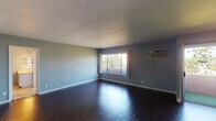 Located at 102 S Oxford Ave in the desirable Hancock Park neighborhood of Los Angeles, this single-floor unit offers a well-organized layout with distinct living, dining, kitchen, and bedroom spaces. Rich, dark hardwood flooring runs throughout the main living areas, creating a striking contrast against the soft blue-gray painted walls and crisp white baseboards and ceilings.

The open living room is generously proportioned, featuring a wall-mounted air conditioning unit and a large window that frames views of the surrounding greenery, filling the space with natural light. A sliding glass door connects the dining area to the exterior, while an elegant multi-arm brass chandelier adds a decorative focal point overhead. The kitchen is designed in a galley style with floor-to-ceiling dark espresso cabinetry, light countertops, subway tile backsplash, tile flooring, and black appliances including a range and microwave.

The bedroom offers a calm, neutral atmosphere with warm carpeting, white walls, and a bright window fitted with horizontal blinds. The bathroom is equipped with a dual-sink vanity featuring a yellow countertop, Hollywood-style lighted mirrors, tile flooring, and built-in storage cabinetry, providing both functionality and a retro-inspired character.