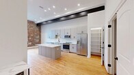 Located at 179 Chelsea Ave in Long Branch, NJ, this striking 3-bedroom, 2-bathroom residence masterfully combines historic character with contemporary design across a single, thoughtfully laid-out floor. Warm hardwood floors flow throughout the space, complemented by soaring ceilings with bold exposed black steel beams that reinforce the industrial aesthetic.

The open-concept kitchen is a standout, featuring sleek gray cabinetry, quartz countertops, a central island, and a full suite of stainless steel appliances including a dishwasher, range, microwave, and refrigerator. Exposed brick walls anchor the kitchen and living areas, adding texture and warmth to the modern palette.

The bedrooms are generously proportioned, with the primary bedroom offering ample space for comfortable living. Several rooms showcase dramatic arched windows set within exposed brick walls, flooding the interior with natural light and adding architectural distinction. A dedicated closet provides additional storage convenience.

Both bathrooms are elegantly finished with marble-style tile surrounds, glass-enclosed showers, and modern black hardware accents. One bathroom features a walk-in shower with mosaic tile flooring, while the second includes a soaking tub combination. The hallway connects all rooms with the same rich hardwood underfoot, creating a cohesive and polished living environment throughout.