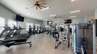 This fitness center is designed to support a comprehensive workout experience across a generously sized, single-level layout. The space is divided into distinct functional zones, each tailored to a specific type of exercise. The main area houses a robust selection of strength-training cable machines and multi-use weight stations, complemented by a full rack of dumbbells positioned in front of a wall-length mirror. A dedicated water fountain station and flat-screen televisions mounted throughout the space add convenience and entertainment during workouts.

The cardio zone features multiple treadmills, elliptical trainers, and a rowing machine, all positioned near large windows that invite natural light and outdoor views. Ceiling fans provide additional air circulation throughout the main floor, enhancing comfort during intense sessions.

A separate room serves as a functional training and stretching area, equipped with stability balls, a vertical knee raise station, and additional free weights, surrounded by multiple windows for a bright, airy atmosphere. French doors in one section of the facility provide direct access to an outdoor area. The neutral-toned tile flooring, soft gray walls, and recessed and linear lighting create a clean, modern ambiance throughout.