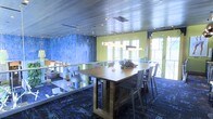 This two-floor space makes a strong visual impression through its inventive use of color, materials, and light. The interior showcases an eye-catching combination of vivid chartreuse-green accent walls, deep blue textured feature walls, and richly patterned blue-toned carpet flooring that adds depth and energy to the environment. A metallic ceiling with recessed lighting lends an industrial yet polished character to the space, reflecting light in a way that enhances the overall ambiance.

A central wooden communal table, surrounded by contemporary metal chairs, serves as a focal point for collaboration and meeting, finished with a suspended linear pendant light fixture above. Glass partitions divide areas while maintaining an open, transparent feel that encourages connectivity between spaces. Natural light filters in through generous windows along the perimeter, brightening the interior and offering views to the outside.

The layout across two floors provides clear separation between functional zones, with visible workstations and collaborative areas visible beyond the glass dividers on the lower level. The overall design balances creativity with functionality, making this a well-appointed space suited for a professional, creative, or collaborative work environment.