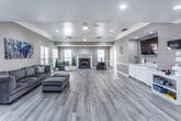 This single-story clubhouse in Forrest City, Arkansas, offers a generously proportioned open-plan interior designed for community gatherings and social activities. The main gathering area is defined by light gray walls, crisp white crown molding, and recessed lighting complemented by dual ceiling fans, creating a bright and welcoming atmosphere throughout.

Light wood-tone luxury vinyl plank flooring extends across the entire space, reinforcing the clean, contemporary aesthetic. A central fireplace with a white-painted surround and blue tile hearth serves as a natural focal point, flanked by windows fitted with white plantation shutters that allow ample natural light while maintaining privacy.

The seating area accommodates comfortable sectional sofas and a coffee table arranged around the fireplace, providing an inviting space for residents to gather. On one side of the room, built-in white cabinetry with a countertop and sink offers a convenient service area, accompanied by a wall-mounted television for entertainment.

A separate zone near the entrance features a glass-top desk and open shelving, suitable for administrative or office use. Multiple windows and glass-panel entry doors along the perimeter further enhance the sense of openness, offering views of the surrounding exterior while maintaining a bright, airy ambiance throughout the clubhouse.
