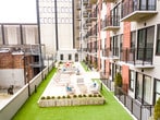This expansive rooftop terrace is designed for outdoor entertaining and relaxation, offering a well-appointed amenity space with sweeping views of the surrounding urban skyline. The terrace is finished with large-format porcelain tile pavers that define the main walkways and gathering areas, complemented by sections of artificial turf that add a touch of greenery and visual contrast throughout the space.

At the heart of the terrace sits a fully equipped outdoor kitchen station featuring dual stainless steel grills set into a stone-finished built-in counter, ideal for al fresco dining and social gatherings. Multiple weathered wood dining tables paired with vibrant red and white contemporary chairs are arranged throughout, providing ample seating for groups of various sizes.

String lights are strung overhead across the length of the terrace, creating a warm and inviting ambiance during evening hours. Raised planter boxes filled with lush white flowering shrubs add natural beauty along the perimeter. A life-sized outdoor chess set on the turf provides a fun recreational element, while lounge seating areas offer spots to unwind.

The terrace is bordered by the building's modern multi-story façade, featuring a mix of concrete, warm wood cladding, large glass windows, and private balconies. The open-air setting delivers unobstructed city views and a bright, airy atmosphere throughout the day.