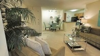 This single-floor residence offers a thoughtfully designed open-concept layout that seamlessly connects the main living areas. The living room is anchored by a comfortable neutral-toned sofa accented with decorative pillows, complemented by a sleek glass coffee table and lush indoor tropical plants that bring a refreshing touch of greenery to the space. Soft, wall-to-wall carpeting in a warm beige tone flows throughout, creating a cohesive and inviting atmosphere. The dining area sits just beyond the living space, featuring a round glass-top table surrounded by elegant teal upholstered chairs, illuminated by a stylish track lighting fixture overhead. A circular wall mirror adds a decorative focal point to the dining space. The kitchen is partially open to the dining area via a raised counter that serves as a natural dividing element, offering views into the kitchen where warm wood-toned cabinetry and modern appliances are visible. The space is bathed in warm, ambient lighting that enhances the comfortable and well-appointed feel of the interior. A front entry door with clean white trim is visible from the main living area, adding to the functional flow of the floor plan. Overall, the space presents a balanced blend of comfort, style, and practical design.