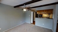 This single-floor space presents an open-concept layout that seamlessly connects the living area to the kitchen. The living space is defined by its most striking architectural feature: dark, exposed wood ceiling beams that run across the ceiling, adding a rustic character to the otherwise neutral interior. A ceiling fan with wood-toned blades is centrally mounted, providing both function and visual interest beneath the beams.

The living area is finished with wall-to-wall carpeting in a light, neutral tone, complementing the cool grey-blue walls throughout. The open sightlines create a comfortable and airy feel within the space.

Adjacent to the living area, the kitchen is partially separated by a raised bar-height counter with a dark stone or laminate surface, offering an informal dining or prep area. The kitchen is equipped with warm-toned wood cabinetry, a built-in microwave, and a stainless range, providing functional cooking amenities. A refrigerator is visible at the kitchen's edge. Beyond the kitchen, a hallway leads to additional areas of the unit, finished with hardwood-style flooring that contrasts the carpeted living space.