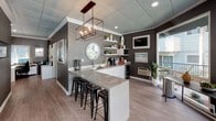 Located at 425 Merrimac Way in Costa Mesa, California, this single-story community clubhouse offers a thoughtfully designed gathering space with a contemporary coastal aesthetic. The interior is anchored by a central kitchen bar featuring a quartz countertop, undermount sink, and sleek white cabinetry, complemented by an eye-catching geometric bronze pendant light overhead. Black metal barstools line the island, creating a casual seating option ideal for socializing. A dedicated refreshment station along the back wall includes open floating shelves styled with coastal décor, a coffee maker, and a beverage refrigerator — all set against a richly textured dark gray accent wall. The dining area is bathed in natural light streaming through expansive floor-to-ceiling windows and glass doors that frame views of the outdoor courtyard, pool area, and mature palm trees beyond. Light wood-look flooring runs throughout, enhancing the bright and airy atmosphere. A wall-mounted TV adds a functional entertainment element to the lounge corner. The tiled ceiling with recessed lighting and pendant fixtures lends a polished finish to the space, while the overall design balances modern sophistication with a relaxed, community-friendly ambiance perfect for residents to gather and connect.
