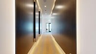 This thoughtfully designed single-floor amenity suite offers a sophisticated blend of collaborative and private workspaces across a generous footprint. The space is anchored by a sleek, illuminated corridor clad in rich wood-toned wall panels with warm cove lighting above and a boldly patterned carpet runner below, setting an elevated tone from entry.

The main gathering area features a large conference table surrounded by mid-century modern seating, a striking geometric chandelier, and a coffered ceiling with ambient cove lighting. Floor-to-ceiling windows flood the room with natural light and offer open views of the surrounding landscape, including palm tree-lined streetscapes. A vibrant lounge area nearby provides comfortable high-back chairs in warm amber tones paired with teal ottomans, creating an inviting environment for informal meetings or relaxation.

Two enclosed private work pods, accessible through navy-framed glass doors, offer focused work settings with individual desks, windows, and colorful accent walls. A dedicated conference room features a bold yellow graphic accent wall, built-in display screen, and integrated power at the table. Patterned modular carpeting with geometric motifs ties the various zones together cohesively. The overall ambiance is contemporary and professional, with a vibrant color palette and abundant natural light throughout.