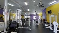 This fitness center offers a generously sized, open workout floor designed to accommodate a wide range of strength and conditioning exercises. The space is immediately recognizable by its bold color scheme, featuring vibrant purple brick accent walls paired with bright yellow highlights, creating an energetic and motivating atmosphere for exercise.

The workout area is furnished with a solid selection of equipment, including cable machines, free weight stations with barbells and weight plates, and adjustable benches, providing users with the tools needed for full-body strength training. A large wall-mounted mirror runs along one side of the space, allowing members to monitor their form during workouts while also enhancing the sense of openness throughout the room.

The ceiling features multiple ceiling fans alongside fluorescent lighting fixtures, ensuring the space remains well-ventilated and brightly lit at all times. The dark rubberized flooring provides both durability and safety underfoot, making it well-suited for heavy equipment use.

Overall, this fitness center delivers a functional and vibrant environment built for serious training, with a practical layout that maximizes the use of available floor space while maintaining clear pathways between equipment stations.