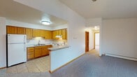 This single-floor residence offers a thoughtfully designed layout with a warm, functional aesthetic defined by consistent wood-toned trim, doors, and cabinetry throughout. The kitchen is a practical centerpiece, featuring an L-shaped arrangement of honey-toned oak cabinets, light granite-style countertops, tile flooring, a double sink, and a full suite of white appliances including a refrigerator, electric range, and dishwasher. A peninsula counter connects the kitchen to the adjacent living area, which boasts vaulted ceilings, plush carpeting, a ceiling fan, and a chandelier-lit dining nook. A separate living room offers a corner fireplace, baseboard heating, and a large sliding glass door that fills the space with natural light and provides access to an outdoor patio. The bedrooms are carpeted and bright, each featuring wood-framed windows that invite natural light, with one bedroom offering direct access to a private bathroom. The bathroom is finished with tile flooring, a vanity with under-sink storage, a lighted mirror, and a tub/shower combination. Throughout the home, consistent baseboard heating, warm neutral walls, and cohesive wood accents create a comfortable and inviting atmosphere.