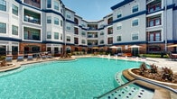 This three-story residential complex is defined by its striking exterior design, blending classic brick accents with crisp white facades, bold navy blue trim, and vibrant red entry doors that frame private balconies on each floor. The building wraps around a generous central courtyard in a sweeping curved configuration, creating an intimate yet open-air atmosphere filled with natural sunlight throughout the day.

At the heart of the complex lies a resort-style swimming pool with clear turquoise water, decorative tiled accents, and thoughtfully landscaped island features. The surrounding pool deck is finished in large-format natural stone pavers, providing an upscale and durable surface for outdoor living. Lounge chairs with colorful cushions, market umbrellas, and outdoor dining sets are arranged across the deck, offering ample space for relaxation and social gatherings.

A dedicated cabana or pavilion structure is visible at one end of the pool area, providing shaded seating and a central gathering point. Manicured hedgerows, tropical palms, and seasonal plantings soften the hardscape and add lush greenery throughout the courtyard. Iron fencing defines the perimeter of the outdoor space while maintaining an open and connected feel. The overall ambiance is that of a well-appointed, amenity-rich community designed for comfortable outdoor living.