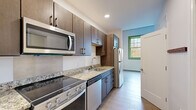This thoughtfully designed single-floor unit offers a clean, modern aesthetic highlighted by light wood-tone flooring and crisp white walls throughout. The well-appointed kitchen is a standout feature, boasting rich dark-wood cabinetry, granite-look countertops, and a full suite of stainless steel appliances including a slide-in electric range, over-the-range microwave, dishwasher, and refrigerator. Ample upper and lower cabinet storage ensures generous organization space, while green-trimmed windows in the adjacent area invite natural light and views of the outdoors. The open living area flows seamlessly from the kitchen, providing a bright, versatile space with light hardwood floors and clean sightlines. The bedroom is notably airy, featuring soaring ceilings that lend a sense of volume and openness, along with additional green-framed windows that reinforce the connection to the outside. The bathroom is designed with accessibility in mind, offering a wide-format tiled floor, a dark wood vanity with an integrated white sink, a large wall mirror, and multiple stainless steel grab bars for added safety and convenience. Recessed lighting and a neutral color palette tie the entire unit together in a cohesive, modern style.