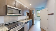 This thoughtfully designed single-floor unit offers a clean, modern aesthetic highlighted by light wood-tone flooring and crisp white walls throughout. The well-appointed kitchen is a standout feature, boasting rich dark-wood cabinetry, granite-look countertops, and a full suite of stainless steel appliances including a slide-in electric range, over-the-range microwave, dishwasher, and refrigerator. Ample upper and lower cabinet storage ensures generous organization space, while green-trimmed windows in the adjacent area invite natural light and views of the outdoors. The open living area flows seamlessly from the kitchen, providing a bright, versatile space with light hardwood floors and clean sightlines. The bedroom is notably airy, featuring soaring ceilings that lend a sense of volume and openness, along with additional green-framed windows that reinforce the connection to the outside. The bathroom is designed with accessibility in mind, offering a wide-format tiled floor, a dark wood vanity with an integrated white sink, a large wall mirror, and multiple stainless steel grab bars for added safety and convenience. Recessed lighting and a neutral color palette tie the entire unit together in a cohesive, modern style.