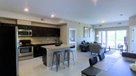 This single-floor space showcases an open-concept design that seamlessly connects the kitchen, dining, and living areas. The kitchen is outfitted with dark espresso cabinetry extending to the ceiling, providing ample storage, while stainless steel appliances, including a range and microwave, add a modern touch. A granite-topped kitchen island with seating for multiple bar stools serves as both a functional workspace and a casual dining spot. Light tile flooring runs throughout the main living area, contributing to a clean, cohesive aesthetic. The living space is bright and inviting, benefiting from natural light streaming in through a sliding glass door or large window, which also offers an outdoor view. Neutral wall tones complement the contemporary furnishings. A doorway visible from the main area leads directly into a bedroom, suggesting a practical and efficient floor plan. Recessed lighting and additional ceiling fixtures provide well-distributed illumination throughout. The overall ambiance is modern and functional, with a thoughtful balance of style and practicality.