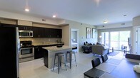 This single-floor space showcases an open-concept design that seamlessly connects the kitchen, dining, and living areas. The kitchen is outfitted with dark espresso cabinetry extending to the ceiling, providing ample storage, while stainless steel appliances, including a range and microwave, add a modern touch. A granite-topped kitchen island with seating for multiple bar stools serves as both a functional workspace and a casual dining spot. Light tile flooring runs throughout the main living area, contributing to a clean, cohesive aesthetic. The living space is bright and inviting, benefiting from natural light streaming in through a sliding glass door or large window, which also offers an outdoor view. Neutral wall tones complement the contemporary furnishings. A doorway visible from the main area leads directly into a bedroom, suggesting a practical and efficient floor plan. Recessed lighting and additional ceiling fixtures provide well-distributed illumination throughout. The overall ambiance is modern and functional, with a thoughtful balance of style and practicality.