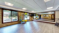 Located at 200 E Burnsville Pkwy in Burnsville, Minnesota, this yoga and fitness studio offers a well-appointed, single-floor space designed to support a variety of exercise and wellness activities. The open studio floor is finished with smooth, hardwood-style flooring that provides an ideal surface for yoga, stretching, and group fitness classes. A large wall-mounted mirror spans a significant portion of the room, enhancing the sense of space and allowing practitioners to monitor their form during workouts. The studio is naturally lit by a generous set of large windows along the exposed brick wall, offering views of the surrounding outdoor greenery and creating a bright, energizing atmosphere. The drop-panel ceiling features recessed lighting to ensure even illumination throughout the space. Equipment is thoughtfully organized within the studio, including a rack stocked with stability balls in various sizes, resistance bands, medicine balls, exercise mats, and a set of colorful dumbbells. A wall-mounted flat-screen display is also available for guided instruction. The entryway area features warm-toned walls accented with framed landscape artwork, wood-trimmed doors, and glass panel entry doors, creating a welcoming transition into the active studio space. This well-equipped studio is suited for yoga, Pilates, group fitness, and personal training sessions.