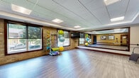 Located at 200 E Burnsville Pkwy in Burnsville, Minnesota, this yoga and fitness studio offers a well-appointed, single-floor space designed to support a variety of exercise and wellness activities. The open studio floor is finished with smooth, hardwood-style flooring that provides an ideal surface for yoga, stretching, and group fitness classes. A large wall-mounted mirror spans a significant portion of the room, enhancing the sense of space and allowing practitioners to monitor their form during workouts. The studio is naturally lit by a generous set of large windows along the exposed brick wall, offering views of the surrounding outdoor greenery and creating a bright, energizing atmosphere. The drop-panel ceiling features recessed lighting to ensure even illumination throughout the space. Equipment is thoughtfully organized within the studio, including a rack stocked with stability balls in various sizes, resistance bands, medicine balls, exercise mats, and a set of colorful dumbbells. A wall-mounted flat-screen display is also available for guided instruction. The entryway area features warm-toned walls accented with framed landscape artwork, wood-trimmed doors, and glass panel entry doors, creating a welcoming transition into the active studio space. This well-equipped studio is suited for yoga, Pilates, group fitness, and personal training sessions.