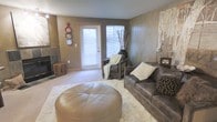This single-floor space centers around a well-appointed living area designed with a warm, earthy color palette of taupes, grays, and browns that creates an inviting and cohesive atmosphere. The room features carpeted flooring partially covered by a plush white shag area rug, which anchors the seating arrangement and adds a layer of texture and comfort underfoot. A rich leather sofa paired with a matching armchair forms the heart of the sitting area, complemented by a large round leather ottoman that serves as both a focal point and functional centerpiece. Natural light filters into the space through a glazed exterior door fitted with horizontal blinds, brightening the room while maintaining privacy. A corner fireplace with a dark stone surround adds architectural interest and a cozy focal point, while rustic wooden accent pieces, including a natural wood stump side table, reinforce the space's organic aesthetic. Decorative elements such as wall-mounted plate art, woven textile wall hangings, and a tall vase with dried branches contribute to the eclectic yet harmonious character of the room. The overall ambiance is relaxed and layered, blending rustic textures with contemporary furnishings for a lived-in, welcoming feel.
