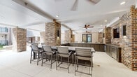 This generously sized amenity space seamlessly blends indoor comfort with outdoor living through a thoughtfully designed covered pavilion and adjacent open-air courtyard. Distinctive exposed brick columns anchor the architecture throughout, providing a warm, textural contrast against crisp white ceilings and neutral-toned wall paneling. The covered area features recessed lighting and multiple ceiling fans, ensuring year-round comfort. A built-in outdoor kitchen serves as a central hub, complete with a beverage cooler, stainless steel refrigerator drawers, dark countertops, and a wall-mounted flat-screen television framed by striking brick walls — ideal for entertaining. High-top bar seating lines one side of the space, while a separate lounge area offers wicker and upholstered seating arrangements for relaxed gatherings. Large-format tile flooring extends throughout, adding a polished, cohesive finish. Opening to the outdoors, the space connects directly to a landscaped courtyard enclosed by a multi-story curved residential building. The courtyard features manicured turf, tropical plantings, umbrella-shaded dining tables, and lounge furniture, creating an inviting, resort-style retreat bathed in natural light.