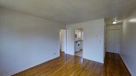 This single-floor space showcases a clean, straightforward layout designed for functional everyday living. The main living area is finished with warm, honey-toned hardwood floors that add character and a sense of warmth throughout. Crisp white walls keep the space feeling bright and neutral, providing a blank canvas for personalization.

Two doorways lead from the main living area — one opening to a hallway that connects to additional rooms, and another providing direct access to the kitchen. The kitchen is compact yet well-organized, featuring white cabinetry, a white gas range, and a countertop workspace. A window in the kitchen allows natural light to filter in, brightening the cooking area.

The layout flows naturally between spaces, making the unit feel cohesive despite its modest scale. The consistent hardwood flooring runs throughout, tying each area together visually. White baseboards trim the walls cleanly, complementing the overall neutral palette. The space offers a simple, well-maintained interior that provides a practical and livable environment.