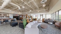 Located at 29 St. Helens Avenue in downtown Tacoma, WA, this thoughtfully designed amenity space blends industrial architecture with contemporary furnishings to create a welcoming and functional environment. The expansive main lounge is anchored by soaring exposed ceilings with white-painted structural trusses, visible ductwork, and a series of decorative globe pendant lights that add warmth and visual interest. Patterned carpet in soft gray tones grounds the space, which is furnished with a generous sectional sofa, accent chairs, and casual seating arrangements suited for both relaxed gatherings and informal meetings. Large windows flood the area with natural light and frame views of the surrounding streetscape. A windowed lounge corridor features floor-to-ceiling glass along one wall, offering sweeping panoramic views of Commencement Bay, the Tacoma waterfront, and the city below — an exceptional vantage point outfitted with plush lounge chairs and a bench for comfortable viewing. The fully equipped kitchen features sleek white cabinetry, dark countertops, stainless steel appliances including a range with hood, dishwasher, and refrigerator, alongside open wood box shelving for a modern, functional aesthetic. A private dining room rounds out the space, highlighted by warm bamboo flooring, a solid wood dining table with leather upholstered chairs, a sculptural ring chandelier, and curated wall art.