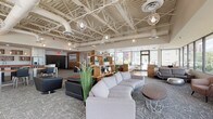 Located at 29 St. Helens Avenue in downtown Tacoma, WA, this thoughtfully designed amenity space blends industrial architecture with contemporary furnishings to create a welcoming and functional environment. The expansive main lounge is anchored by soaring exposed ceilings with white-painted structural trusses, visible ductwork, and a series of decorative globe pendant lights that add warmth and visual interest. Patterned carpet in soft gray tones grounds the space, which is furnished with a generous sectional sofa, accent chairs, and casual seating arrangements suited for both relaxed gatherings and informal meetings. Large windows flood the area with natural light and frame views of the surrounding streetscape. A windowed lounge corridor features floor-to-ceiling glass along one wall, offering sweeping panoramic views of Commencement Bay, the Tacoma waterfront, and the city below — an exceptional vantage point outfitted with plush lounge chairs and a bench for comfortable viewing. The fully equipped kitchen features sleek white cabinetry, dark countertops, stainless steel appliances including a range with hood, dishwasher, and refrigerator, alongside open wood box shelving for a modern, functional aesthetic. A private dining room rounds out the space, highlighted by warm bamboo flooring, a solid wood dining table with leather upholstered chairs, a sculptural ring chandelier, and curated wall art.