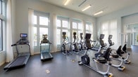 This comprehensive fitness center is designed to support a full range of workout needs across multiple functional zones. The main cardio area is lined with floor-to-ceiling windows dressed in sheer curtains, flooding the space with natural light and offering outdoor views. This area houses a lineup of treadmills, ellipticals, and stationary bikes, all equipped with individual screens for an engaging workout experience.

The strength training zone features a broad selection of cable machines, weight racks, and a full dumbbell set displayed along a wall-length mirror, maximizing both functionality and the sense of space. Mounted televisions throughout keep users informed and entertained during their sessions.

A dedicated group exercise studio provides an open, airy environment with soaring ceilings, sheer-curtained windows, and durable rubberized flooring. The studio is outfitted with a TRX suspension training rig, wall-mounted resistance bands, exercise mats, stability balls, and foam rollers — accommodating yoga, stretching, and functional fitness classes alike.

Adjacent to the main fitness floor, a cheerful play room offers a welcoming retreat, featuring colorful patterned carpet tiles, vibrant wall art, a television, and shelving — ideal for younger visitors. The overall aesthetic throughout is clean and modern, with soft blue-gray walls, recessed and linear lighting, and a cohesive, energizing atmosphere.
