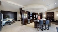 This contemporary cybercafe is thoughtfully designed to blend functionality with modern aesthetics. The single-floor layout features an open, welcoming interior finished with light tile flooring and a neutral ceiling palette that contrasts elegantly with the rich, dark wood cabinetry and architectural column accents throughout the space.

At the heart of the room, a curved white-topped bar counter with sleek bar seating provides a comfortable spot for guests to work or unwind. The cabinetry along the back wall is well-appointed, housing built-in appliances including a compact refrigerator, along with display shelving and digital information screens integrated seamlessly into the design.

Track lighting runs along a curved ceiling rail, casting focused illumination across the workspace and countertops, enhancing the polished, professional ambiance. A vending machine adds a convenient self-service element to the space. An adjacent lounge area is visible beyond the main counter, offering additional seating for those seeking a more relaxed setting.

Overall, the cybercafe presents a clean, upscale atmosphere suited for productivity and leisure alike, with modern finishes and a well-organized layout that ensures ease of use for all visitors.