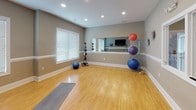 Located at 410 Starmount Park Blvd in Kannapolis, NC, this single-floor fitness and yoga studio is thoughtfully designed to support a wide range of wellness activities. The space features warm hardwood-style flooring throughout, providing a clean and functional surface ideal for yoga, stretching, and floor-based workouts. Neutral gray walls accented with crisp white chair rail molding and baseboards create a calm, focused atmosphere.

A large wall-mounted mirror spans one side of the room, enhancing the sense of space and allowing for form checks during exercise. A flat-screen television is mounted on a dedicated accent wall, enabling guided workout sessions or entertainment during training. Recessed lighting illuminates the room evenly, while windows fitted with white blinds allow for natural light control.

The studio is well-equipped with a medicine ball storage rack holding a full set of weighted balls, multiple stability and exercise balls of varying sizes, and a yoga mat. A pass-through window connects the studio to an adjacent fitness equipment room, offering convenient access to additional machines. Decorative wall accents add a subtle touch of personality to the space. This studio serves as a versatile, well-appointed environment for fitness, yoga, and group or individual wellness programming.