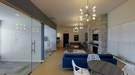 This single-floor communal space is thoughtfully designed to balance comfort and social interaction. The main lounge area is anchored by a striking floor-to-ceiling stone fireplace fitted with a mounted flat-screen television, serving as a natural focal point for the room. Two distinctive sputnik-style chandeliers hang overhead, casting warm ambient light and contributing to the space's contemporary aesthetic.

Seating is arranged to encourage conversation, with a navy blue sectional sofa paired with a light-toned area rug and a central coffee table. Additional modern accent chairs and a small round dining table with sculptural white chairs provide flexible seating options throughout.

Toward the rear of the space, a foosball table adds a recreational element, making this area well-suited for casual gatherings. A built-in workstation or bar counter along the back wall offers a functional surface for additional use.

A glazed, frameless glass-walled room adjacent to the main lounge provides a semi-private enclosure, visible from the common area. Light wood-toned flooring flows throughout, tying the space together with a clean, modern finish.