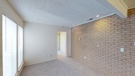 This single-floor residence offers a cohesive and functional layout with notable architectural character throughout. The living area serves as the heart of the space, anchored by a full-length exposed brick accent wall that adds warmth and texture to the room. Soft carpet underfoot, recessed lighting overhead, and a textured ceiling complete the comfortable atmosphere, while an open passage connects the living area directly to the kitchen.

The kitchen is well-appointed with white cabinetry, dark countertops, a contrasting dark tile backsplash, and hardwood-style flooring. White appliances — including a range, dishwasher, and refrigerator — are integrated seamlessly, and a double-basin sink sits beneath convenient counter space, making the kitchen both practical and clean in design.

The bedroom is bright and inviting, featuring plush carpet, a ceiling fan with integrated lighting, a window with blinds for natural light, and a sliding door with vertical blinds that opens to additional space. A generously sized walk-in closet accompanies the bedroom, outfitted with built-in shelving and double hanging rods to maximize storage.

The bathroom includes a wide vanity with dark countertop, dual mirrors with sconce lighting, and a full tub-shower combination, offering a clean and functional design throughout.