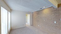 This single-floor residence offers a cohesive and functional layout with notable architectural character throughout. The living area serves as the heart of the space, anchored by a full-length exposed brick accent wall that adds warmth and texture to the room. Soft carpet underfoot, recessed lighting overhead, and a textured ceiling complete the comfortable atmosphere, while an open passage connects the living area directly to the kitchen.

The kitchen is well-appointed with white cabinetry, dark countertops, a contrasting dark tile backsplash, and hardwood-style flooring. White appliances — including a range, dishwasher, and refrigerator — are integrated seamlessly, and a double-basin sink sits beneath convenient counter space, making the kitchen both practical and clean in design.

The bedroom is bright and inviting, featuring plush carpet, a ceiling fan with integrated lighting, a window with blinds for natural light, and a sliding door with vertical blinds that opens to additional space. A generously sized walk-in closet accompanies the bedroom, outfitted with built-in shelving and double hanging rods to maximize storage.

The bathroom includes a wide vanity with dark countertop, dual mirrors with sconce lighting, and a full tub-shower combination, offering a clean and functional design throughout.