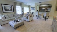 This single-floor space offers a thoughtfully designed open-concept layout that seamlessly connects the living, dining, and kitchen areas into one cohesive and functional environment. The expansive living area features plush carpeting, generous seating arrangements, and large windows dressed with neutral-toned curtains that allow abundant natural light to fill the room, creating a warm and inviting atmosphere. A tufted ottoman serves as a central accent piece, complementing the neutral color palette throughout. The dining area flows naturally from the living space, providing a comfortable spot for gatherings and everyday meals. An elegant arched passageway leads into the kitchen, which is outfitted with dark wood cabinetry, stainless steel appliances, and a raised breakfast bar with high-back seating — ideal for casual dining or entertaining. The kitchen's layout promotes easy interaction with guests in the adjoining living and dining areas. Track lighting and recessed fixtures illuminate the space effectively, while the consistent use of soft, neutral tones across the walls, flooring, and furnishings contributes to an overall ambiance that feels both spacious and refined. The cohesive design and open flow make this a highly functional and welcoming space.