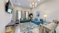 Located at 82 Ardmore Gardens Dr in Hardeeville, SC, this single-floor community space offers a welcoming and well-appointed environment designed for both relaxation and productivity. The main lounge area immediately impresses with its soaring ceilings and an abundance of natural light streaming through multiple windows dressed in elegant patterned drapery. A gold chandelier serves as a striking focal point overhead, complementing the coastal-inspired color palette of soft blues, creams, and warm neutrals throughout.

The seating areas are generously furnished with plush teal sofas, a curved sectional, and patterned accent chairs, all arranged around vibrant botanical and coral-motif area rugs that anchor each conversation space. Original artwork adorns the walls, adding a curated, gallery-like quality to the interiors. Light tile flooring runs throughout, enhancing the bright, airy atmosphere.

A dedicated business center is seamlessly integrated into the space, featuring an L-shaped granite countertop workstation equipped with multiple computers, a multifunction printer, and stylish table lamps — ideal for residents needing a professional workspace. A wall-mounted flat-screen TV provides an additional media amenity within the lounge. The thoughtful layout and cohesive design make this a versatile and inviting community hub.