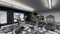 Located at 3701 Governors Club Blvd in Columbus, Ohio, this fitness center offers a well-appointed, single-floor workout space designed to support an active lifestyle. The room features a clean, modern aesthetic with navy blue accent walls contrasted by light-toned trim and a patterned multi-color carpet that adds a contemporary touch underfoot. Bright fluorescent lighting runs along the ceiling, complementing the natural light that flows in through multiple windows fitted with blinds, providing views of the surrounding greenery. The cardio area is well-equipped with a selection of Precor machines, including upright and recumbent stationary bikes, treadmills, and elliptical trainers, arranged to allow comfortable spacing between users. Wall-mounted flat-screen televisions are positioned for easy viewing from multiple workout stations, enhancing the exercise experience. The space maintains a functional and organized layout, making it accessible and easy to navigate. Overall, the fitness center delivers a focused and motivating environment suited for both casual and routine exercise within the Governors Club community.