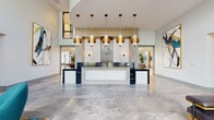 Located at 7200 N Frontage Rd in Orlando, FL, this single-floor amenity center is a showcase of contemporary design and refined finishes. The grand lobby greets visitors with a sculptural reception desk framed by gold and white pendant lighting, textured accent walls, and large-format stone tile flooring that flows throughout the space. A "Live. Work." branded focal wall sets the tone for the community's modern lifestyle ethos. Adjacent lounge areas feature bold, curated abstract artwork, ambient backlit wall panels, plush seating, and vibrant area rugs that inject color and warmth into the clean, neutral palette. Floor-to-ceiling windows flood these spaces with natural light while offering views of the surrounding landscaping. The kitchen areas are thoughtfully designed with two-tone cabinetry in navy blue and light wood, quartz and stone countertops, a textured wave backsplash, and a full suite of stainless steel appliances — one featuring a galley-style layout and another with an open peninsula and bar seating. The expansive game and social lounge boasts soaring double-height ceilings, oversized windows draped in sheer curtains, a statement billiards table, and eclectic seating arrangements, creating an inviting space for community gatherings and leisure.