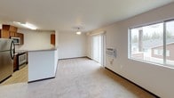 Located at 15001 North Wandermere Road in Spokane, WA, this single-floor unit offers a practical and well-organized layout designed for comfortable everyday living. The open-concept living and kitchen area forms the heart of the space, where a peninsula-style kitchen counter with a light gray laminate surface separates the two zones. The kitchen is equipped with stainless steel appliances — including a refrigerator, microwave, and range — complemented by warm-toned medium brown cabinetry and a double-basin stainless steel sink with ample counter space. Light hardwood-style flooring runs throughout the kitchen, contrasting with the plush neutral carpet in the living area. The living room benefits from a sliding glass door that connects to an outdoor area and is accompanied by a side window, allowing natural light to fill the space. Bedrooms are carpeted and feature windows with views of the surrounding neighborhood and tree line, providing a quiet, comfortable retreat. The bathroom includes a single-sink vanity with warm wood cabinetry, a large mirror with overhead lighting, and hard surface flooring. A floor-level HVAC vent and wall-unit air conditioning ensure year-round climate control throughout the unit.