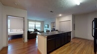 Situated in the heart of downtown Akron, Ohio, this single-floor unit at 401 S Main St offers a well-designed, functional layout ideal for modern living. The open-concept kitchen is a standout feature, centered around a large granite-topped island with bar seating and an integrated sink. Dark espresso cabinetry pairs with black appliances — including a full-size refrigerator, range, and built-in microwave — to create a cohesive, contemporary look. Warm hardwood flooring flows throughout the main living areas, adding a rich, inviting tone to the space. The living room is bright and comfortable, featuring large windows that frame city views of the Akron skyline, providing ample natural light. Multiple bedrooms each include hardwood floors, a built-in desk with wall-mounted shelving, a closet, and windows overlooking the surrounding cityscape — offering a practical setup suited for study or work. The bathrooms are finished with slate-style tile flooring, dark vanity cabinetry with granite countertops, and full tub-shower combinations. The textured ceilings and neutral wall tones throughout lend the unit a clean, understated aesthetic consistent across every room.