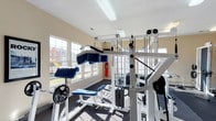 This fitness center offers a comprehensive workout environment designed to support a range of exercise routines. The space is outfitted with a multi-station cable and pulley strength training system, free weights, and a dedicated weight bench, providing users with the tools needed for both upper and lower body conditioning. Rubber-matted flooring covers the workout area, offering both durability and safety underfoot. Large windows line the walls, flooding the interior with natural light and offering views of the surrounding outdoor grounds, including a glimpse of a playground area. The bright, neutral-toned walls and high ceiling contribute to an open and airy atmosphere, making the space feel welcoming and energizing. A fire extinguisher is visibly mounted on the wall, reflecting attention to safety standards. The layout is thoughtfully arranged to allow users to move freely between equipment. Whether focused on strength training or general fitness, this center provides a functional and well-maintained facility suited to a variety of fitness levels and goals.