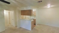 This single-floor residence offers a practical and well-organized layout designed for everyday comfort. The kitchen serves as a central feature of the space, equipped with a white range and oven, a refrigerator, and ample cabinetry in a warm wood finish. Upper cabinetry is prominently positioned above the kitchen zone, providing generous storage capacity. A peninsula-style breakfast bar separates the kitchen from the adjoining living area, creating a natural division between cooking and relaxing spaces while maintaining an open, connected feel throughout. The living area is finished with wall-to-wall carpeting in a neutral tone, contributing to a warm and quiet atmosphere. Walls throughout are painted in a soft, light cream palette, enhancing the sense of brightness in the space. A textured ceiling adds a classic residential character. Overhead HVAC vents are integrated into the ceiling, ensuring climate comfort throughout. The overall layout flows efficiently, making good use of the available footprint. The design is straightforward and functional, offering a clean canvas suitable for a variety of furnishing styles and personal touches.