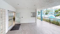 This single-floor space functions as a communal lobby and transitional area, designed with a clean, modern aesthetic. The interior entry zone features smooth concrete flooring, white walls, and recessed ceiling lighting, creating a bright and minimal atmosphere. A glass-panel entry door with a dark entrance mat welcomes occupants, while a glass block window accent wall introduces subtle natural light diffusion without compromising privacy.

The space transitions seamlessly into a semi-covered open-air terrace, distinguished by bold teal-painted structural columns and a low horizontal metal railing. This outdoor-facing area is generously proportioned, offering open views of mature trees, surrounding buildings, and what appears to be a courtyard with a shaded umbrella below. The textured concrete flooring continues throughout, providing a cohesive visual flow between interior and exterior zones.

Two stainless steel elevator doors serve as a focal point near the transition between enclosed and open areas, providing vertical access within the broader building. Security cameras are visibly mounted at key points, indicating a monitored environment. The overall ambiance is functional yet open, with an emphasis on connectivity between indoor comfort and outdoor surroundings.