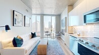 This thoughtfully designed high-rise unit offers a seamless blend of modern industrial aesthetics and contemporary comfort. The open-concept layout connects the living, dining, and kitchen areas into one cohesive space, anchored by light wide-plank hardwood flooring and striking exposed concrete ceilings with integrated track lighting throughout.

Floor-to-ceiling windows dominate the main living area, flooding the space with abundant natural light while framing sweeping panoramic views of the surrounding urban skyline. The living area provides a comfortable and stylish setting, complemented by a compact dining nook positioned directly beside the expansive glazing to take full advantage of the cityscape.

The kitchen is well-appointed with sleek white cabinetry, subway tile backsplash, a stainless steel appliance suite including a gas range, built-in microwave, French door refrigerator, and dishwasher, along with ample counter space for everyday use.

The bathroom is finished to a high standard, featuring large-format light wood-toned tile flooring, a warm wood vanity with an illuminated mirror, and a glass-enclosed walk-in shower. A private entryway hallway provides a clean transition into the main living space. The overall ambiance is bright, airy, and polished, with a modern urban character defined by its elevated position and expansive city views.