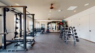This single-story fitness and wellness facility opens into a striking reception lounge distinguished by soaring vaulted ceilings, a statement rope chandelier, and light hardwood flooring. Framed large-scale equestrian artwork flanks a central hallway, while woven lounge chairs arranged around a sculptural wooden coffee table create a welcoming, design-forward entrance. A wall-mounted display screen and glass double doors connect the space to the outdoors, flooding the area with natural light.

The facility encompasses multiple dedicated fitness zones. A spacious strength training room features rubber flooring, a full power rack, extensive free weight racks, TRX suspension systems, a heavy punching bag, and floor-to-ceiling mirrors framed in warm wood trim. An adjacent cardio room offers a bright, window-lined environment equipped with treadmills, ellipticals, and rowing machines, providing views of lush greenery. A separate studio room with light hardwood floors, a wall-mounted TV, and framed mirrors provides an ideal space for yoga, stretching, or low-impact training. A functional training area with stability balls, resistance tools, and ample open floor space rounds out the workout offerings.

A private treatment room, finished in calming grey tones with wood-paneled walls and curated artwork, provides a serene setting for massage and recovery services, completing the full wellness experience.