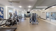This fitness center is a comprehensively outfitted facility designed to accommodate a wide range of workout routines. The space is organized into distinct zones, allowing for efficient use of the open floor plan. The cardio area features multiple treadmills, stationary upright bikes, and spin cycles, providing ample options for aerobic training. A large wall-mounted flat-screen television is positioned centrally, enhancing the workout experience.

The strength training zone is equally well-appointed, housing a multi-station cable machine, a variety of selectorized weight machines branded by Matrix, benches, and a dedicated free weights section complete with a full dumbbell rack. Large mirrors line the walls, amplifying the sense of space and allowing users to monitor their form throughout their workouts.

The facility features durable, easy-to-clean flooring throughout, with ceiling fans providing air circulation alongside recessed and linear LED lighting that ensures the space is consistently bright. Arched windows allow natural light to filter in, adding an airy quality to the environment. A glass-paneled door connects to an adjacent room, offering additional functionality. The overall ambiance is clean, modern, and purpose-built for fitness.
