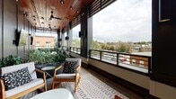 Located on W Huntingdon Street in Savannah, Georgia, this single-level outdoor terrace offers a well-appointed open-air experience elevated above the surrounding neighborhood. The space is designed as a long, covered terrace with a rich wood-planked ceiling that adds warmth and architectural character throughout. String lights and track lighting are strung overhead, providing ambient and functional illumination, while ceiling fans ensure comfortable airflow.

The terrace is divided into distinct functional zones. A lounge area features seating arrangements with accent pillows, a hanging egg chair, and patches of artificial turf adding a touch of greenery and comfort underfoot. Large planter boxes filled with lush greenery are positioned throughout, contributing to the space's inviting atmosphere.

A dedicated dining area includes a long wooden table with sleek black chairs, ideal for group gatherings, while a mounted flat-screen television nearby enhances the entertainment possibilities. Additional seating vignettes with wooden armchairs and patterned area rugs create intimate conversation areas.

The open-railing perimeter provides unobstructed panoramic views of Savannah's low-rise rooftops, mature tree canopy, and distant church steeples, firmly anchoring the terrace within the character of this historic city.