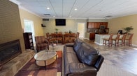 This well-appointed single-floor space is designed for communal gathering and functionality, offering a thoughtfully arranged open-concept layout that seamlessly connects several distinct areas. The lounge area serves as a welcoming focal point, furnished with leather seating and patterned armchairs arranged around a glass-topped coffee table. A striking floor-to-ceiling brick fireplace anchors this zone, flanked by windows with exterior views and a small bookcase, adding warmth and character to the space.

Adjacent to the lounge, a dedicated dining area accommodates a large wooden table with seating for multiple guests, positioned beneath a wall-mounted flat-screen television. The flooring transitions between tile and area rugs, delineating each functional zone clearly.

The kitchen is equipped with medium-toned wood cabinetry, stone tile backsplash, stainless steel appliances including a range and dishwasher, and a multi-seat peninsula counter ideal for casual dining or serving. A separate conference or meeting room features its own large wooden table, ample seating, and a wall-mounted display screen.

Throughout, drop ceilings with recessed lighting and crown molding provide a polished, professional finish. Natural light filters in through multiple windows, creating a bright and inviting atmosphere across the entire space.