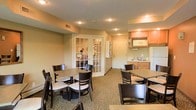 This single-floor space is designed to serve as a communal dining and lounge area, offering a welcoming and functional environment for guests or residents. The main dining area features multiple square tables paired with dark wood chairs with cushioned seats, arranged to accommodate both small and larger groups comfortably. The flooring is covered in a patterned carpet that adds warmth and texture throughout the space.

A compact kitchenette is situated along one wall, equipped with upper cabinetry in a warm wood finish, a microwave, a refrigerator, and a sink with counter space — providing convenient access to light food and beverage preparation. Recessed lighting in the textured ceiling ensures the space is well-lit throughout.

A set of elegant French doors with glass panes leads to an adjoining room, visible through the doors as an office or front desk area, suggesting this space may function as part of a hotel, extended-stay lodging, or residential community facility. The overall color palette of warm beiges and soft greens creates a relaxed, neutral ambiance. The layout is practical and open, designed to encourage casual gathering while maintaining an orderly, tidy atmosphere suitable for everyday use.
