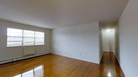 This single-floor space offers a straightforward, functional layout characterized by its clean lines and minimalist design. The main living area is anchored by richly toned hardwood floors that have been polished to a high shine, adding warmth and a sense of quality to the otherwise understated interior. Crisp white walls throughout create a bright, neutral backdrop that lends the space a fresh, move-in-ready feel.

Natural light enters through a window positioned along one wall, providing a view of surrounding greenery and contributing to the airy atmosphere of the room. A baseboard heating unit runs beneath the window, ensuring efficient climate control during cooler months. The ceiling is smooth and white, maintaining a cohesive, uncluttered aesthetic.

A hallway visible toward one end of the space suggests additional rooms beyond the main living area, hinting at a practical and connected floor plan. The overall ambiance is calm and neutral, making it a versatile space well-suited for a variety of furnishing styles and uses. The combination of hardwood flooring, ample wall space, and natural light gives the interior a timeless, adaptable character.