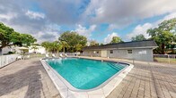Located at 1601 Northwest 13th Street in Boca Raton, Florida, this outdoor pool area offers a refreshing amenity space typical of South Florida living. The centerpiece is a generously sized rectangular swimming pool with clear turquoise water, framed by a well-maintained paver deck that provides ample space for poolside relaxation. Entry into the pool is made easy via built-in steps with metal handrails positioned at the corner of the pool. The surrounding area is enclosed by a metal security fence, ensuring a safe environment. Lounge chairs are visible along the pool's edge, offering spots to enjoy the Florida sunshine. A low-profile utility building adjacent to the pool appears to house restroom or storage facilities, finished in a neutral stucco exterior. The backdrop is enriched by lush, mature tropical landscaping, including palm trees and broad-canopied shade trees that provide a natural, verdant screen. The open sky above allows for abundant natural light, making this an inviting outdoor retreat. The overall ambiance is casual and functional, well-suited to the warm Boca Raton climate.