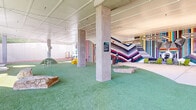 This dynamic, single-level recreational facility is designed to engage and entertain, blending covered indoor spaces seamlessly with open-air outdoor areas. The interior play zone is anchored by concrete columns supporting a clean white paneled ceiling strung with warm string lights, creating an inviting atmosphere. Artificial turf covers much of the floor, complemented by large natural boulder accents that add an organic, playful element to the space. Bold, large-scale murals dominate the interior walls, featuring vibrant chevron and vertical stripe patterns in a rich spectrum of colors, alongside a striking large-format portrait of a French Bulldog — strongly suggesting this is a purpose-built pet amenity facility. Circular pedestal play elements and grass mounds provide interactive features throughout. The outdoor sections extend the experience with expansive artificial turf runs enclosed by horizontal wood privacy fencing and concrete retaining walls, set against a naturally wooded hillside backdrop. Tall light poles illuminate the outdoor areas, and mature trees and landscaped mulch beds line the perimeter. The facility transitions fluidly between its sheltered and open-air zones through wide openings supported by structural columns, ensuring visibility and airflow throughout.