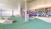 This dynamic, single-level recreational facility is designed to engage and entertain, blending covered indoor spaces seamlessly with open-air outdoor areas. The interior play zone is anchored by concrete columns supporting a clean white paneled ceiling strung with warm string lights, creating an inviting atmosphere. Artificial turf covers much of the floor, complemented by large natural boulder accents that add an organic, playful element to the space. Bold, large-scale murals dominate the interior walls, featuring vibrant chevron and vertical stripe patterns in a rich spectrum of colors, alongside a striking large-format portrait of a French Bulldog — strongly suggesting this is a purpose-built pet amenity facility. Circular pedestal play elements and grass mounds provide interactive features throughout. The outdoor sections extend the experience with expansive artificial turf runs enclosed by horizontal wood privacy fencing and concrete retaining walls, set against a naturally wooded hillside backdrop. Tall light poles illuminate the outdoor areas, and mature trees and landscaped mulch beds line the perimeter. The facility transitions fluidly between its sheltered and open-air zones through wide openings supported by structural columns, ensuring visibility and airflow throughout.